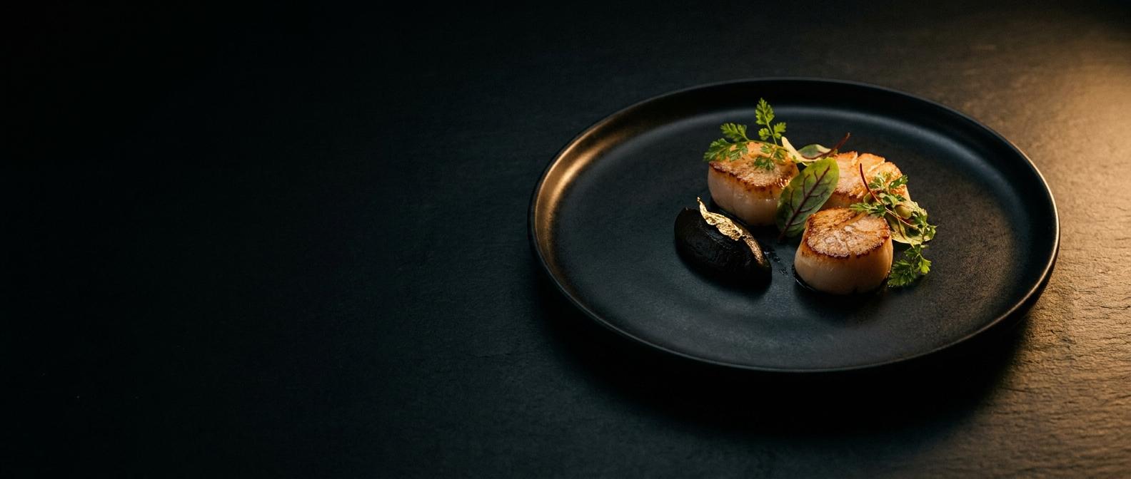 Seared scallops with black garlic purée and gold leaf on a black stoneware plate